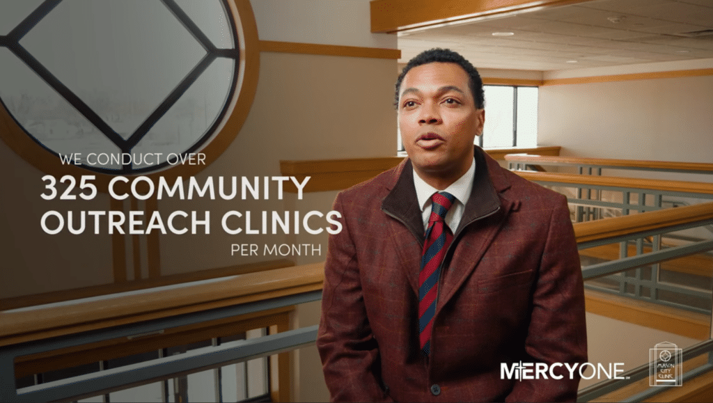 A man in a red blazer and striped tie speaks to the camera. Behind him is a modern office space with large windows. Text on the image reads, "We conduct over 325 community outreach clinics per month." The bottom corner displays "MercyOne" and associated logos.