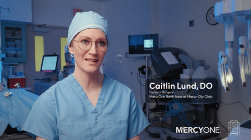 A medical professional in light blue surgical scrubs and a matching cap speaks in a clinical setting with various medical equipment in the background. The text on the image reads: "Caitlin Lund, DO General Surgery MercyOne North Iowa at Mason City Clinic" along with the MercyOne logo.