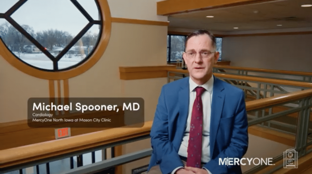 A man in a blue suit and red tie sits in an indoor hallway with wooden railings and large windows. The text on the image reads "Michael Spooner, MD, Cardiology, MercyOne North Iowa at Mason City Clinic." The MercyOne logo is visible in the bottom right corner.