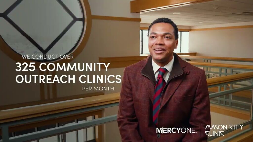 A man in a maroon jacket and striped tie stands inside a building. Text on the image reads, “We conduct over 325 community outreach clinics per month.” Logos for MercyOne and Mason City Clinic are displayed at the bottom. The background shows a modern interior with large windows.
