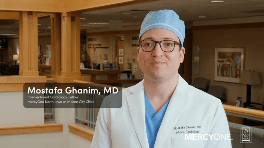 A man wearing glasses, a blue surgical cap, and a white coat stands in a medical facility. The text beside him reads "Mostafa Ghanim, MD, Interventional Cardiology, Fellow" with healthcare logos including MercyOne.