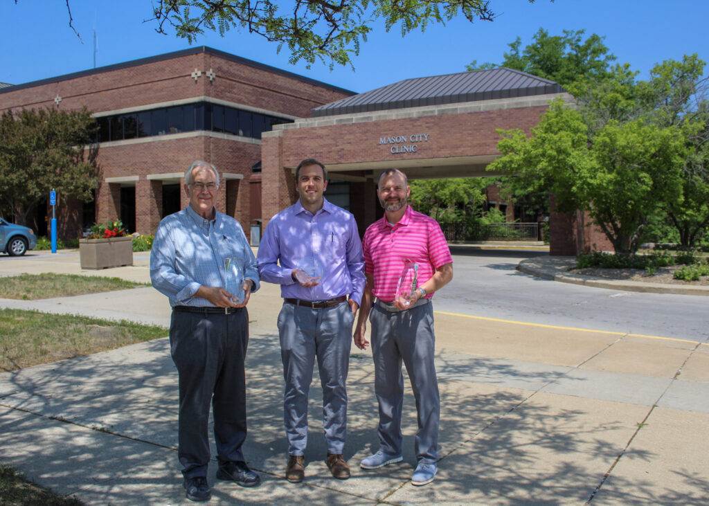 Three men are standing in front of Mason City Clinic, holding glass awards. The clinic is a brick building partially covered by trees, with a visible sign that reads "Mason City Clinic." The men are dressed in business casual attire, and the weather is sunny.