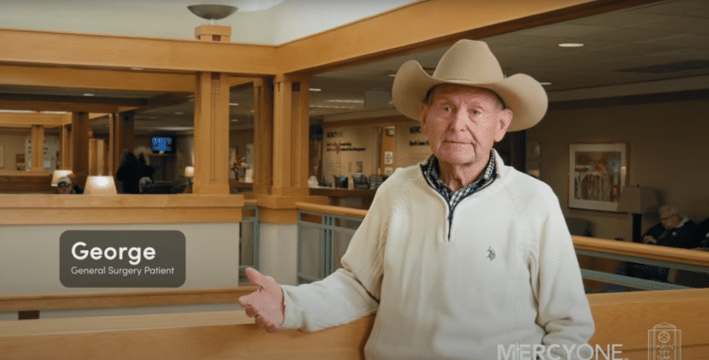 A man wearing a cowboy hat and a white sweater stands in a spacious, warmly lit hallway in a medical facility. A label next to him reads "George - General Surgery Patient." The background shows wooden railings, framed art, and other people seated.