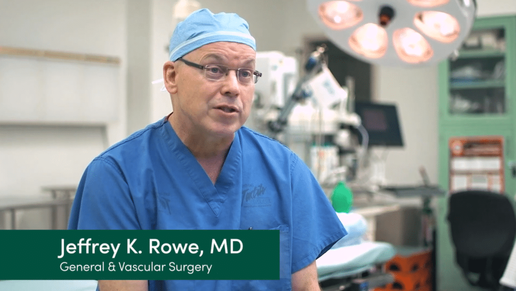 A man wearing blue surgical scrubs and a cap is seated in an operating room setting. A nameplate on the image reads "Jeffrey K. Rowe, MD, General & Vascular Surgery." Medical equipment and surgical lights are visible in the background.