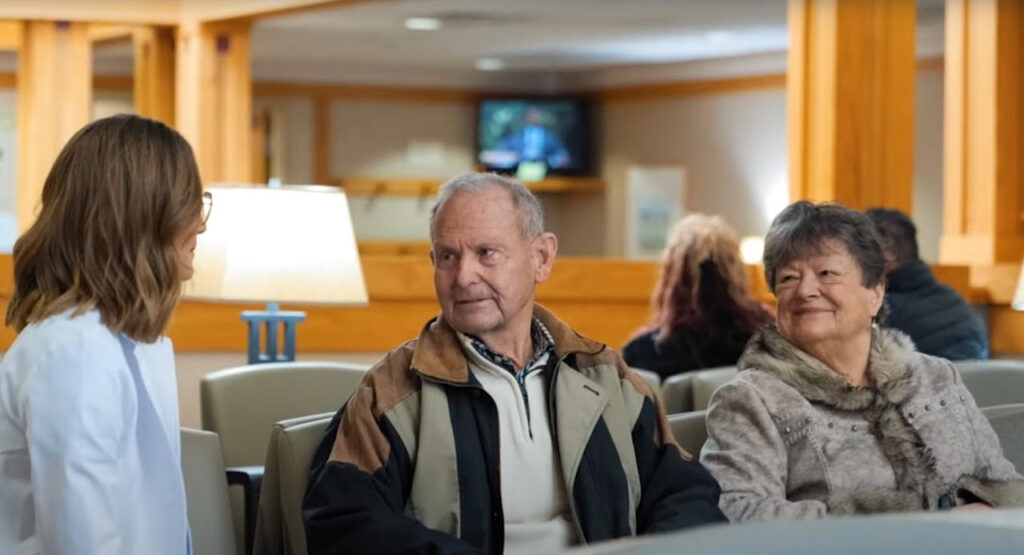 An elderly man and woman sit in a waiting area, engaged in conversation with a woman in a white coat, likely a medical professional. The setting appears to be a clinic or hospital, with people and a television in the background.