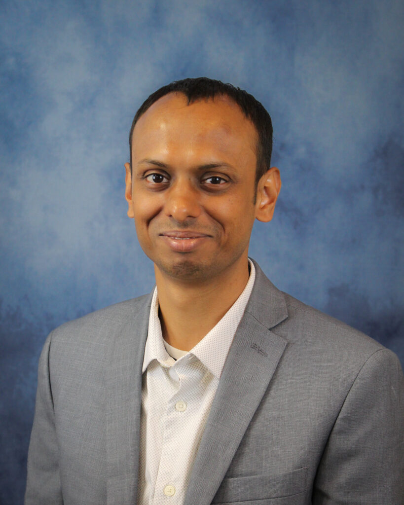 A man with short black hair is smiling for a professional headshot. He is wearing a light gray blazer over a white button-down shirt. The background is a blue and gray mottled pattern.