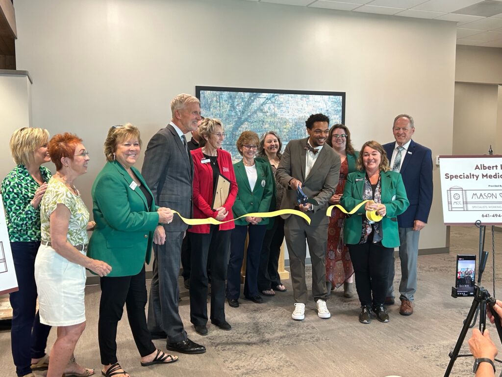 A group of people is gathered as a ribbon is cut during a ceremony. Most individuals are wearing green blazers. A man in a grey suit is cutting the yellow ribbon with large scissors, while others smile and look on. A sign and phone stand are also visible.