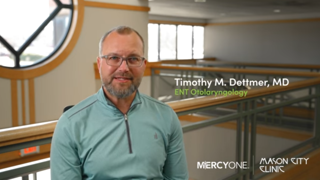 A man in a light blue sweater sits in an office building. The text reads: "Timothy M. Dettmer, MD, ENT Otolaryngology," with logos for MercyOne and Mason City Clinic in the lower corners.