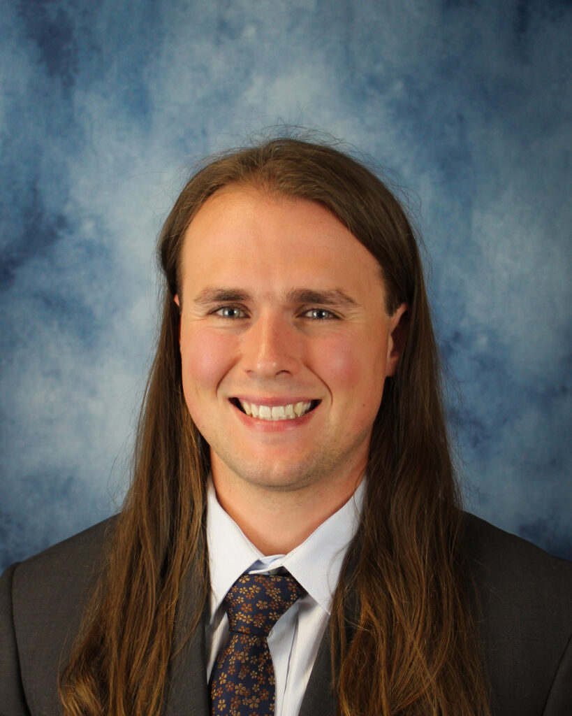 A person with long brown hair, wearing a suit and tie, smiles in front of a blue textured background.