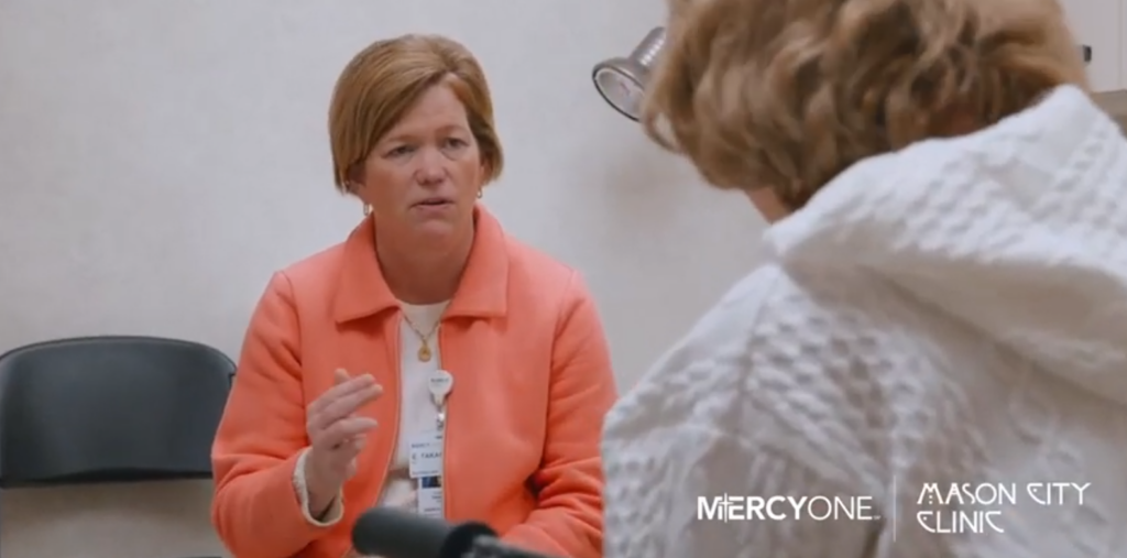 A healthcare professional in an orange jacket gestures while talking to a patient with a white sweater in a clinical setting. Logos for MercyOne and Mason City Clinic are visible on the bottom right.