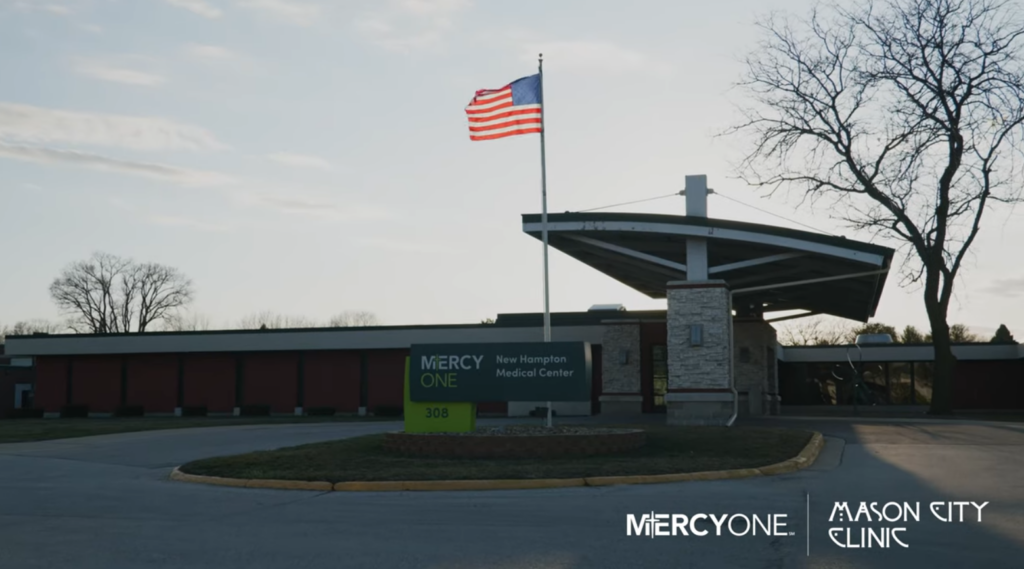 American flag flying at the entrance of MercyOne New Hampton Medical Center, with building and signs for MercyOne and Mason City Clinic in view. Overcast sky in the background.