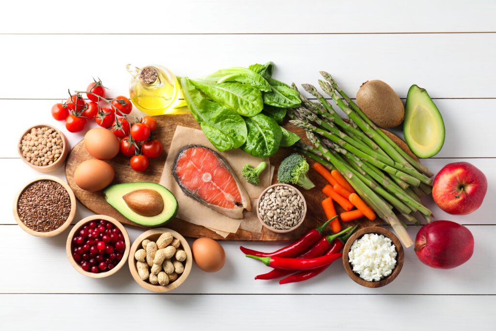 A variety of fresh foods on a table, including salmon, avocado, asparagus, lettuce, cherry tomatoes, carrots, cranberries, nuts, grains, eggs, chili peppers, apples, cottage cheese, and olive oil.