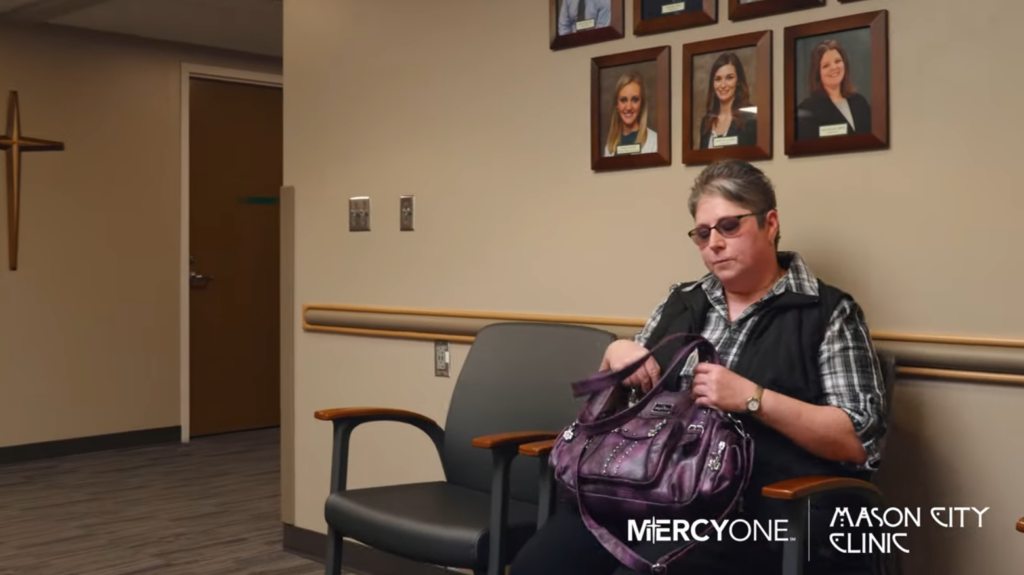 A woman sits alone in a clinic waiting room with beige walls. She's looking into a purple handbag. Behind her are framed photographs of smiling professionals. The room is labeled "MercyOne Mason City Clinic.