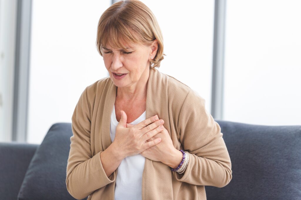 A woman sitting on a couch holds her chest with both hands, appearing to be in discomfort. She is wearing a beige cardigan over a white top. The background consists of large windows.