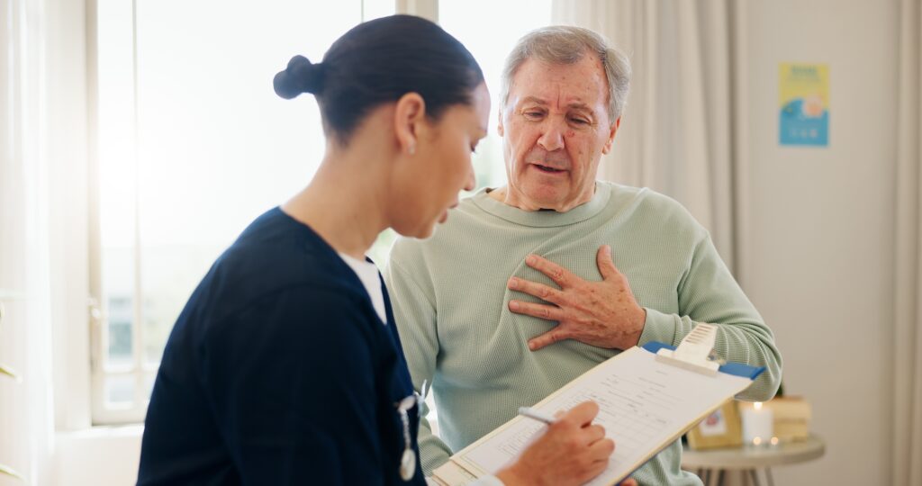 A healthcare professional in a navy uniform holds a clipboard and talks to an elderly man sitting beside her. The man, wearing a light green sweater, gestures with his hand on his chest as they converse in a bright room.