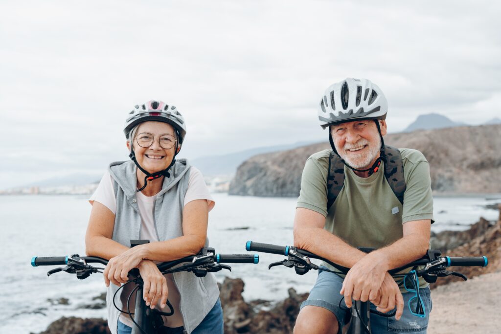 An elderly couple wearing helmets is smiling while leaning on their bicycles near a rocky coastline. The background shows the ocean and distant hills under a cloudy sky.