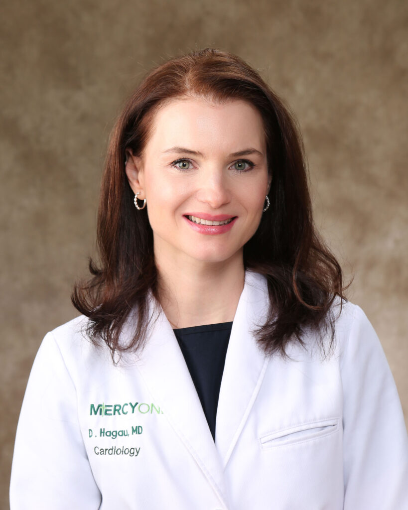 A person with shoulder-length brown hair is smiling at the camera. They are wearing a white lab coat with "MercyOne" and "D. Hagau, MD, Cardiology" embroidered on it. The background is a neutral brown gradient.