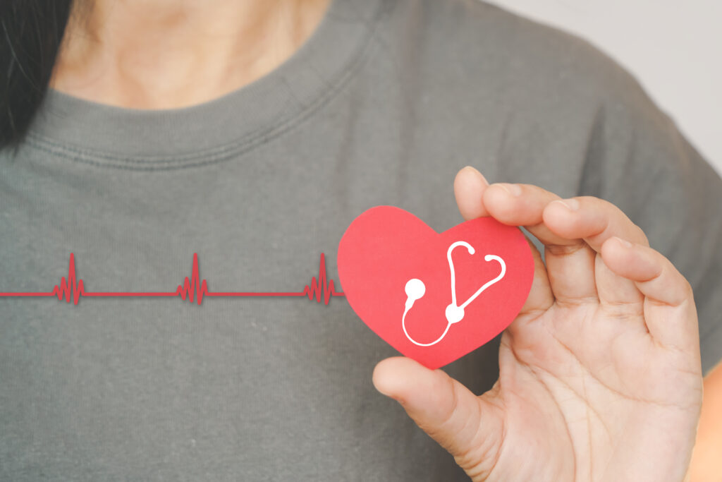 A person holds a red paper heart with a stethoscope symbol, set against a gray shirt. A red heartbeat line runs horizontally across the image.