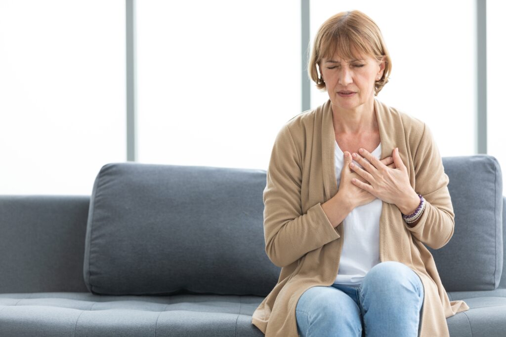 A woman sitting on a couch with her eyes closed and hands on her chest, appearing to experience discomfort. She's wearing a beige cardigan and jeans. The background features a large window with soft natural light.