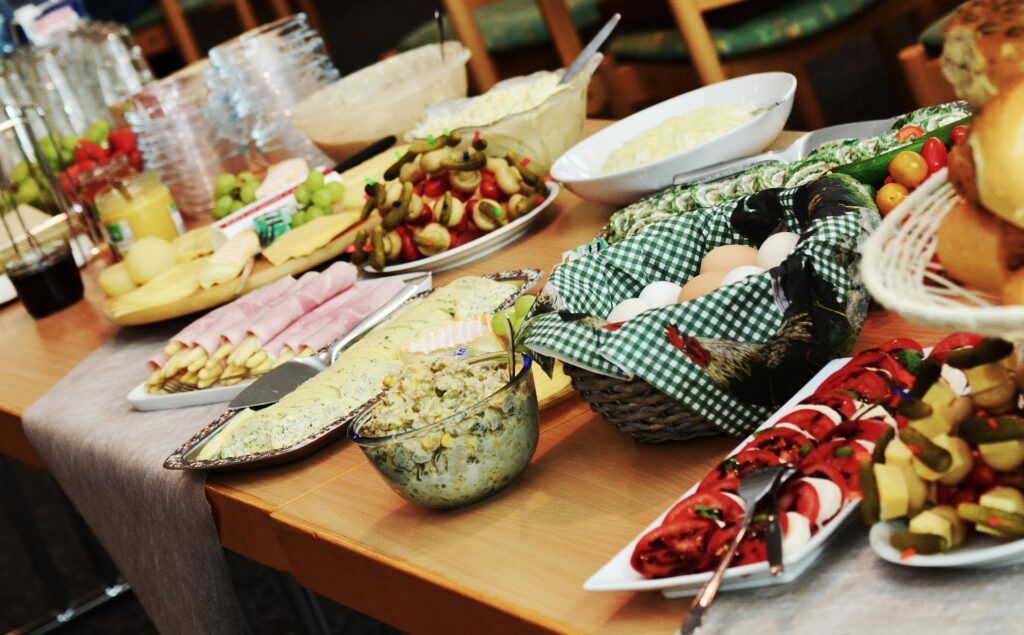 A table laden with an assortment of foods including skewers, sliced ham, hard-boiled eggs, salads, bread rolls, and a variety of cheeses. Plates and cups are neatly arranged nearby, suggesting a buffet-style setup.