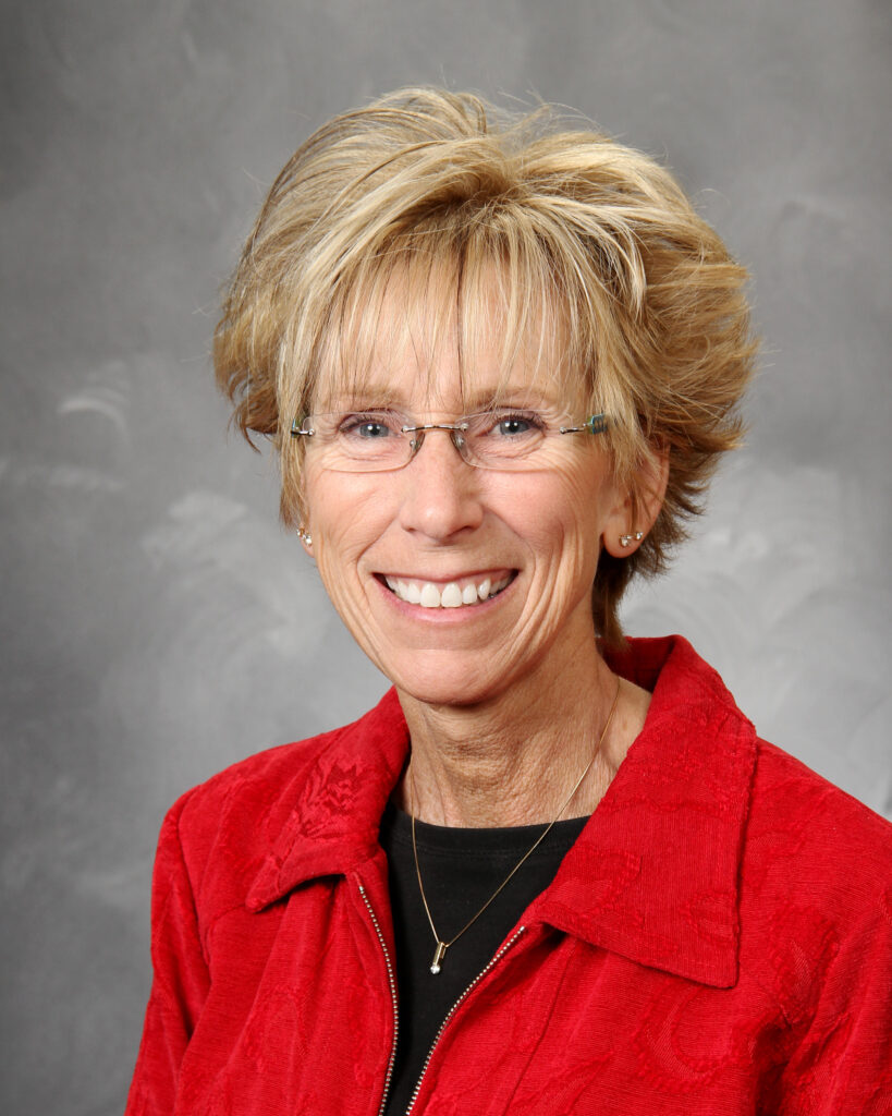 A person with short, light-colored hair, wearing glasses and a red jacket over a black top, smiles at the camera against a gray background.