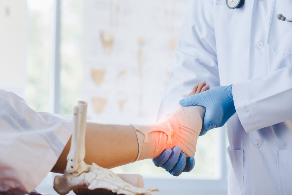 A medical professional in a white coat and blue gloves examines a patient's bandaged foot. The doctor gently supports the foot, while an anatomical model of a leg is partially visible beside them, suggesting the context of a medical setting.