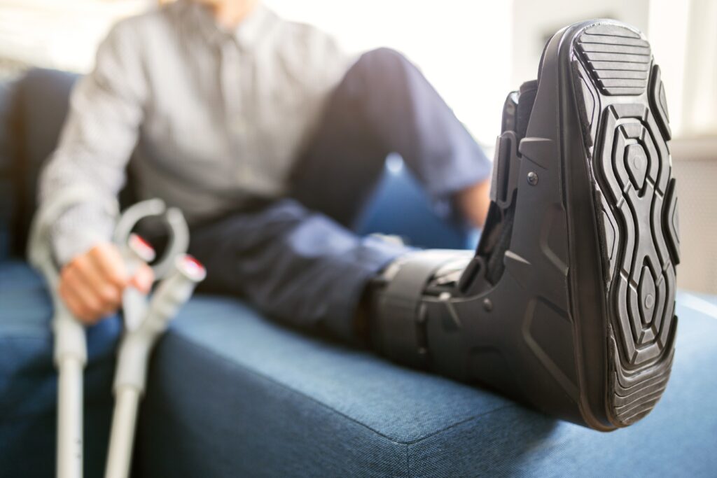 Person sitting on a blue couch with their leg elevated, wearing a black orthopedic boot. They are holding crutches and wearing a checkered shirt and jeans. The focus is on the boot.