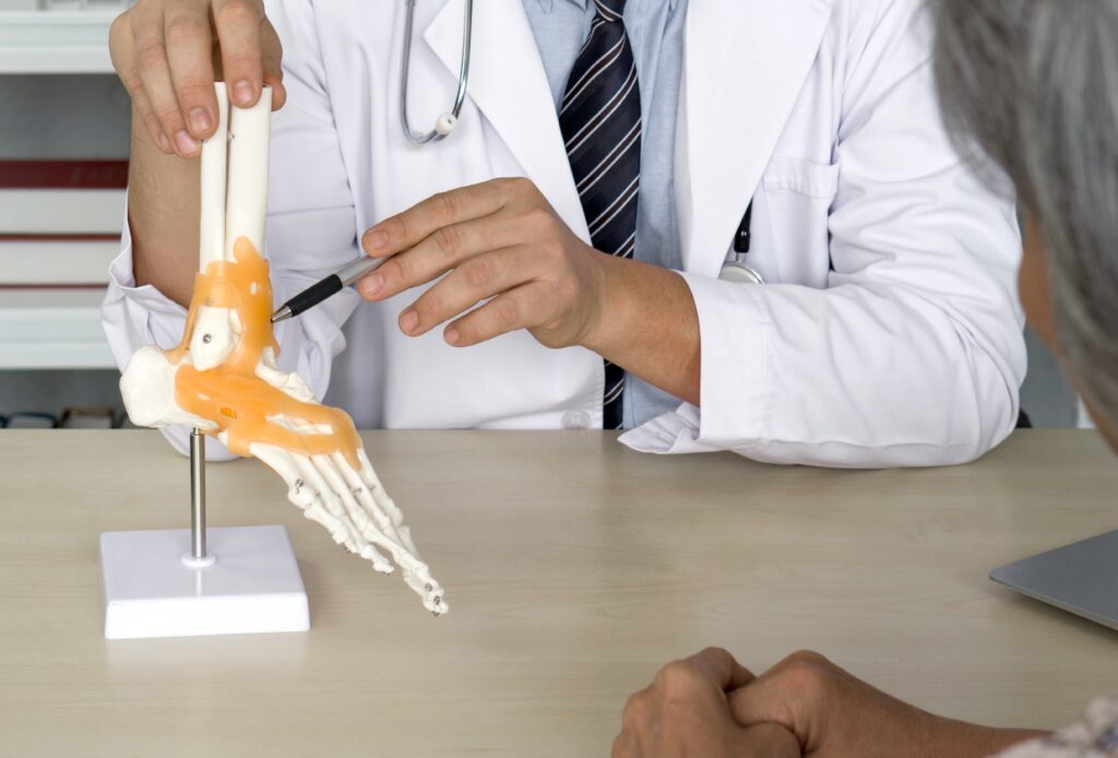 A doctor in a white coat and stethoscope uses a pen to explain a foot and ankle model to a patient sitting across the desk. The model is detailed, showing bones and joints, and the patient listens attentively.