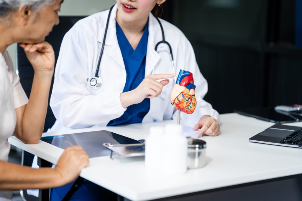 A doctor in a white coat and stethoscope is explaining a heart model to a patient. They are sitting at a desk with medical items and a laptop. The patient appears to be listening attentively.