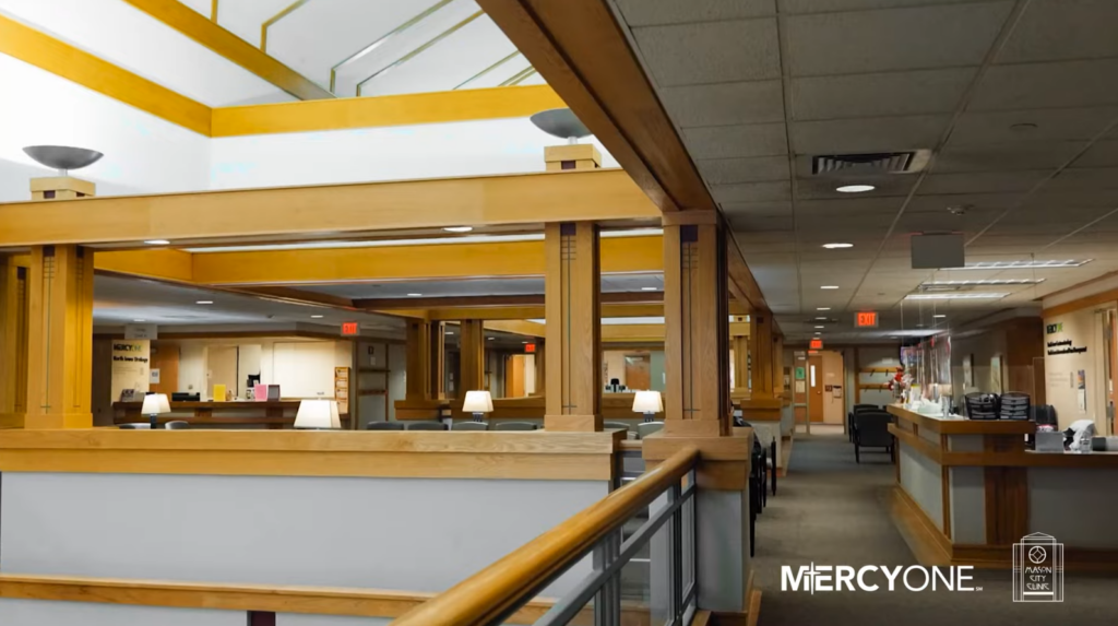 Interior of a building with wooden accents, a railing, and a carpeted hallway leading to a reception area with lit lamps. The ceiling has slanted beams. Logos for "MercyOne" and "Ankeny City Centre" are visible.