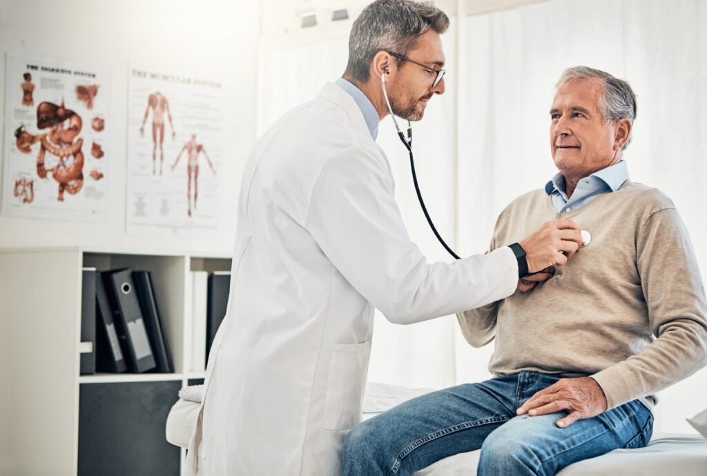 A doctor in a white coat uses a stethoscope to listen to an elderly man's chest in a medical office. The man is seated on an examination table, wearing a gray sweater and jeans. Medical posters are visible on the wall in the background.