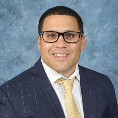 A person wearing glasses, a dark checkered suit, white shirt, and yellow tie is smiling against a textured blue background.