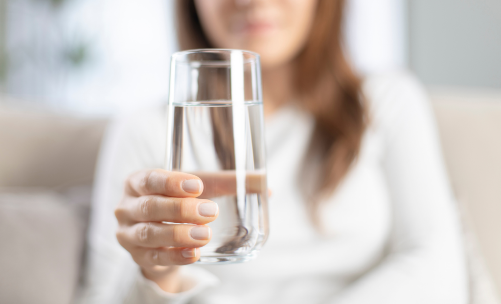 A person with long brown hair, wearing a white shirt, holds a clear glass of water toward the camera. The background is softly blurred.
