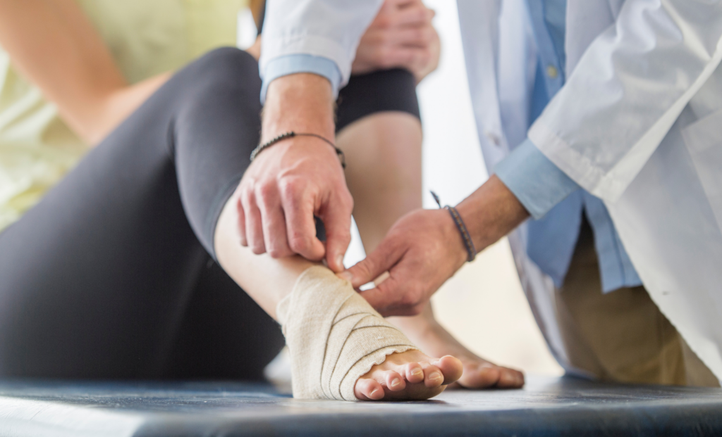 A medical professional wraps a bandage around a person's injured ankle while the person sits with one knee bent on an examination table.