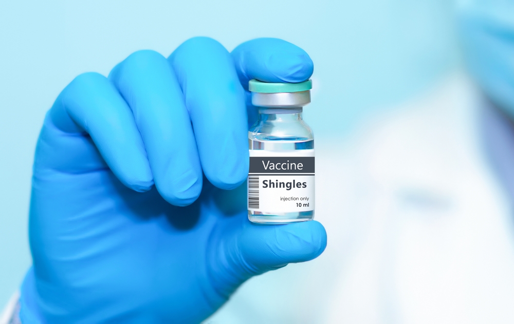 A gloved hand holds a vial labeled "Vaccine Shingles, injection only, 10 ml" against a light blue background.