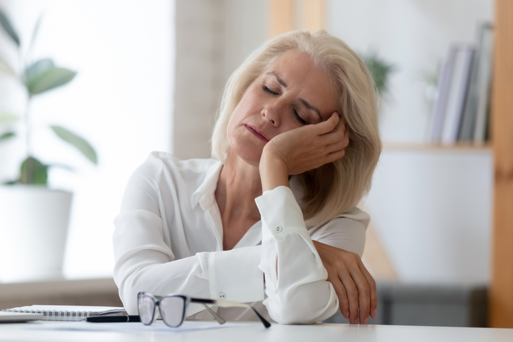 A woman with blonde hair sits at a desk, resting her head on her hand with her eyes closed, appearing tired or sleepy. Glasses and papers are on the table in front of her.