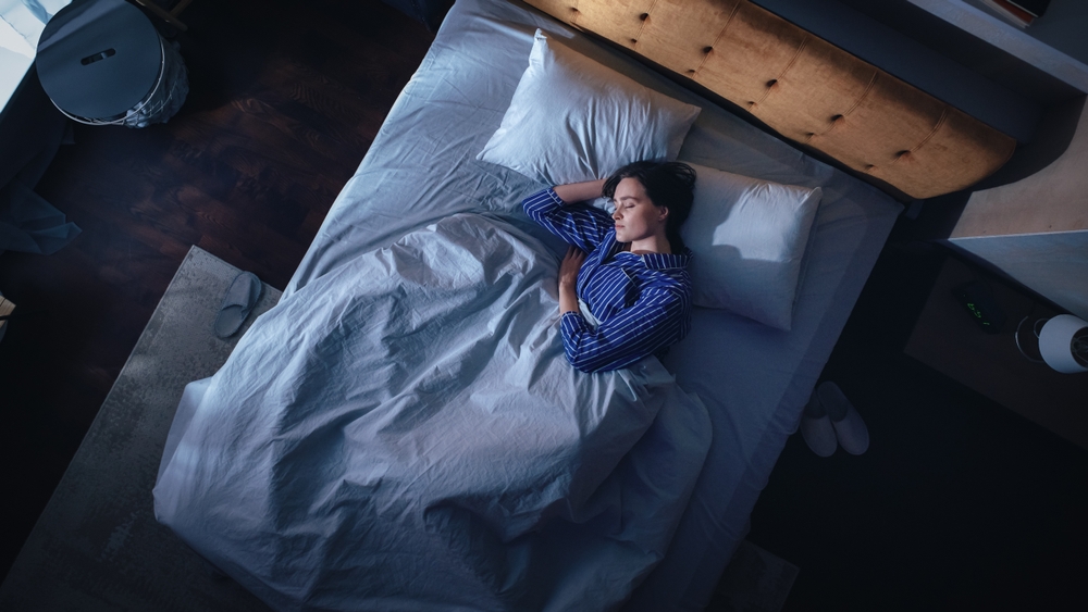 A woman in blue pajamas sleeps on her side in a bed with white sheets and pillows, viewed from above in a dimly lit bedroom at night.