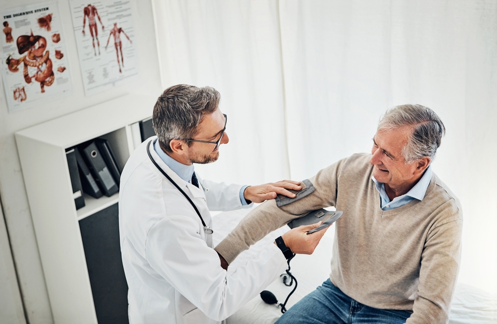 A doctor takes the blood pressure of an older man in a medical office. The patient smiles while sitting on an examination table. Medical charts and folders are visible in the background.