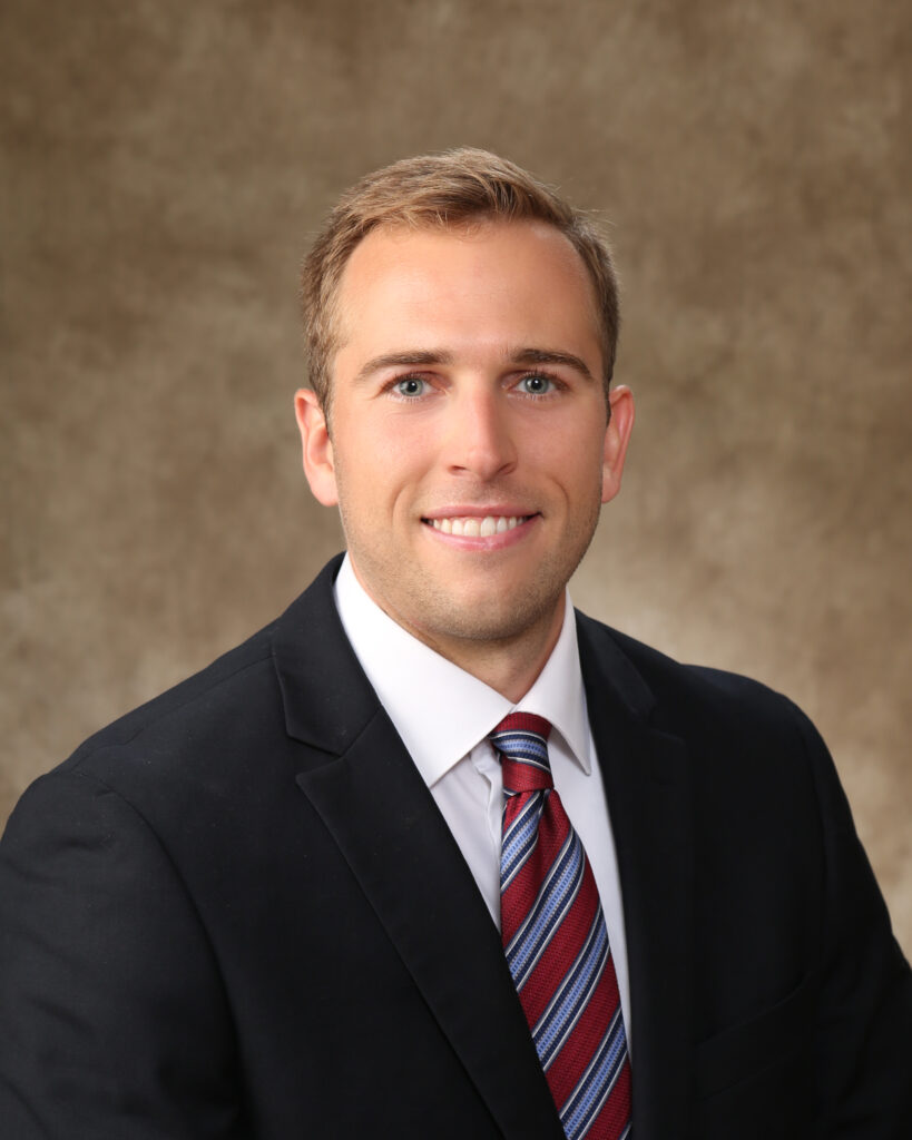 A man with short light brown hair wearing a black suit jacket, white dress shirt, and a red and blue striped tie, smiling in front of a brown textured background.